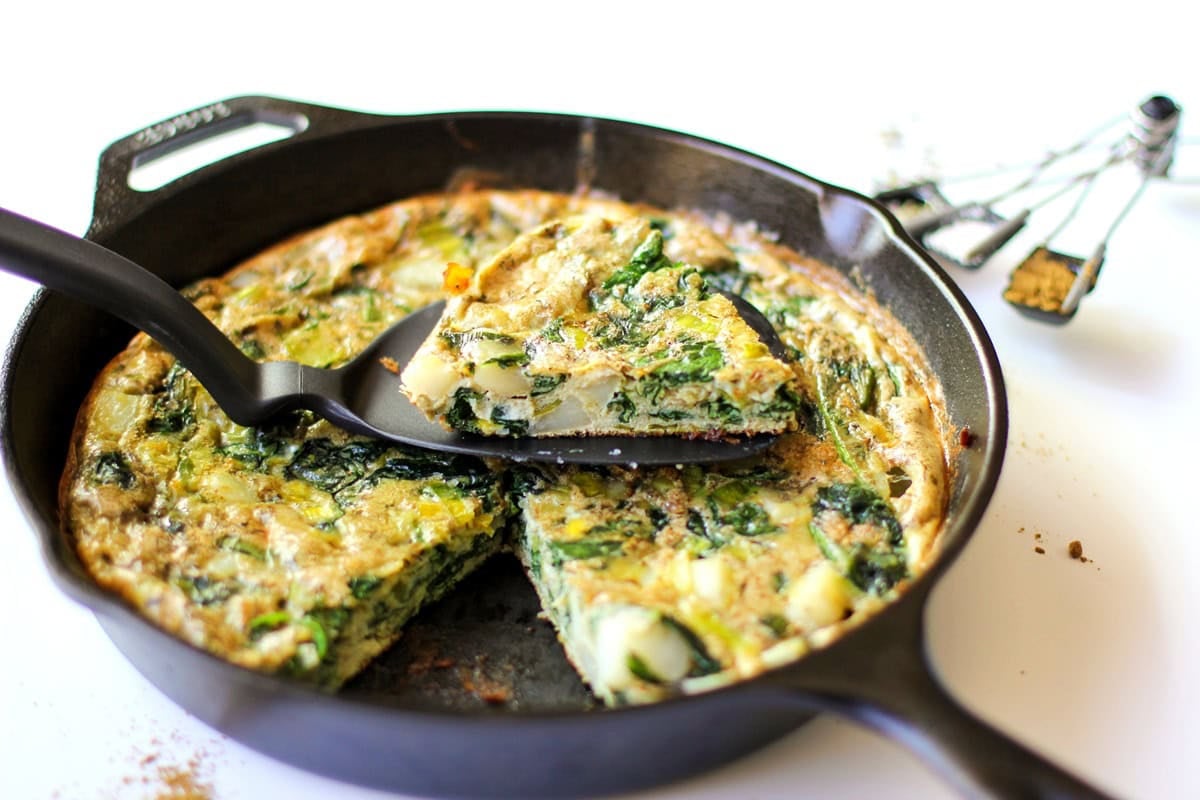 Cast iron skillet full of a frittata with spinach, leeps, and potatoes. Ready to serve with a slice taken out.