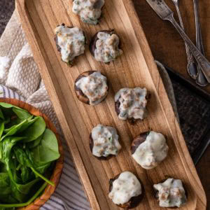 Meat and spinach stuffed mushrooms topped with melted cheese and herbs, arranged on a wooden plate. The baked mushrooms feature a golden-brown color and a savory, flavorful filling.