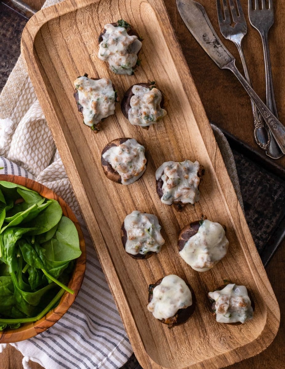 Meat and spinach stuffed mushrooms topped with melted cheese and herbs, arranged on a wooden plate. The baked mushrooms feature a golden-brown color and a savory, flavorful filling.
