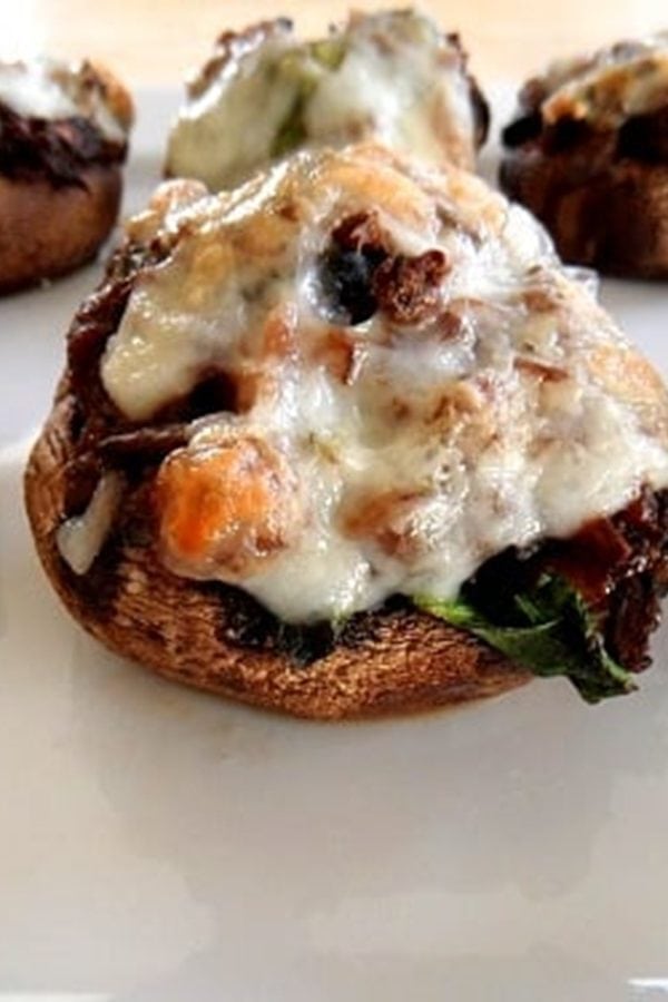 Close-up of meat and spinach stuffed mushrooms topped with melted cheese and herbs, arranged on a white plate. The baked mushrooms feature a golden-brown color and a savory, flavorful filling.