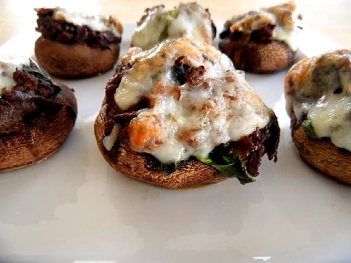 Close-up of meat and spinach stuffed mushrooms topped with melted cheese and herbs, arranged on a white plate. The baked mushrooms feature a golden-brown color and a savory, flavorful filling.