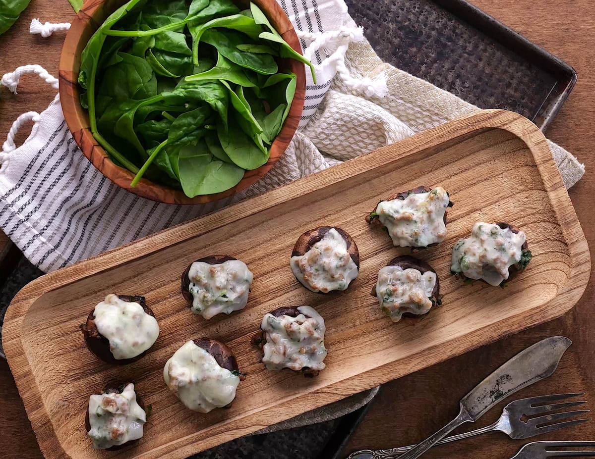 Stuffed mushrooms on a wooden platter on top of a wooden table with silverware to the side, ready to serve.