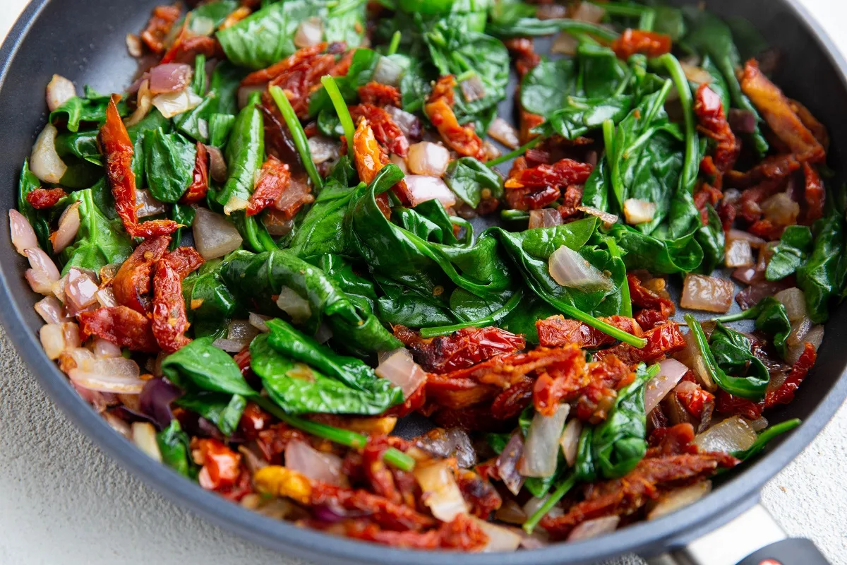 Onions, sun-dried tomatoes and fresh spinach in a sauté pan, to be used as topping for the salmon.