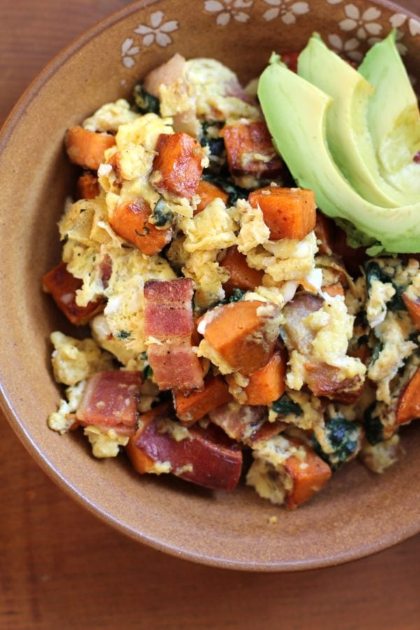 A brown bowl filled with a hearty scramble of eggs, diced sweet potatoes, spinach, and pieces of ham, topped with slices of fresh avocado. The bowl sits on a wooden surface.