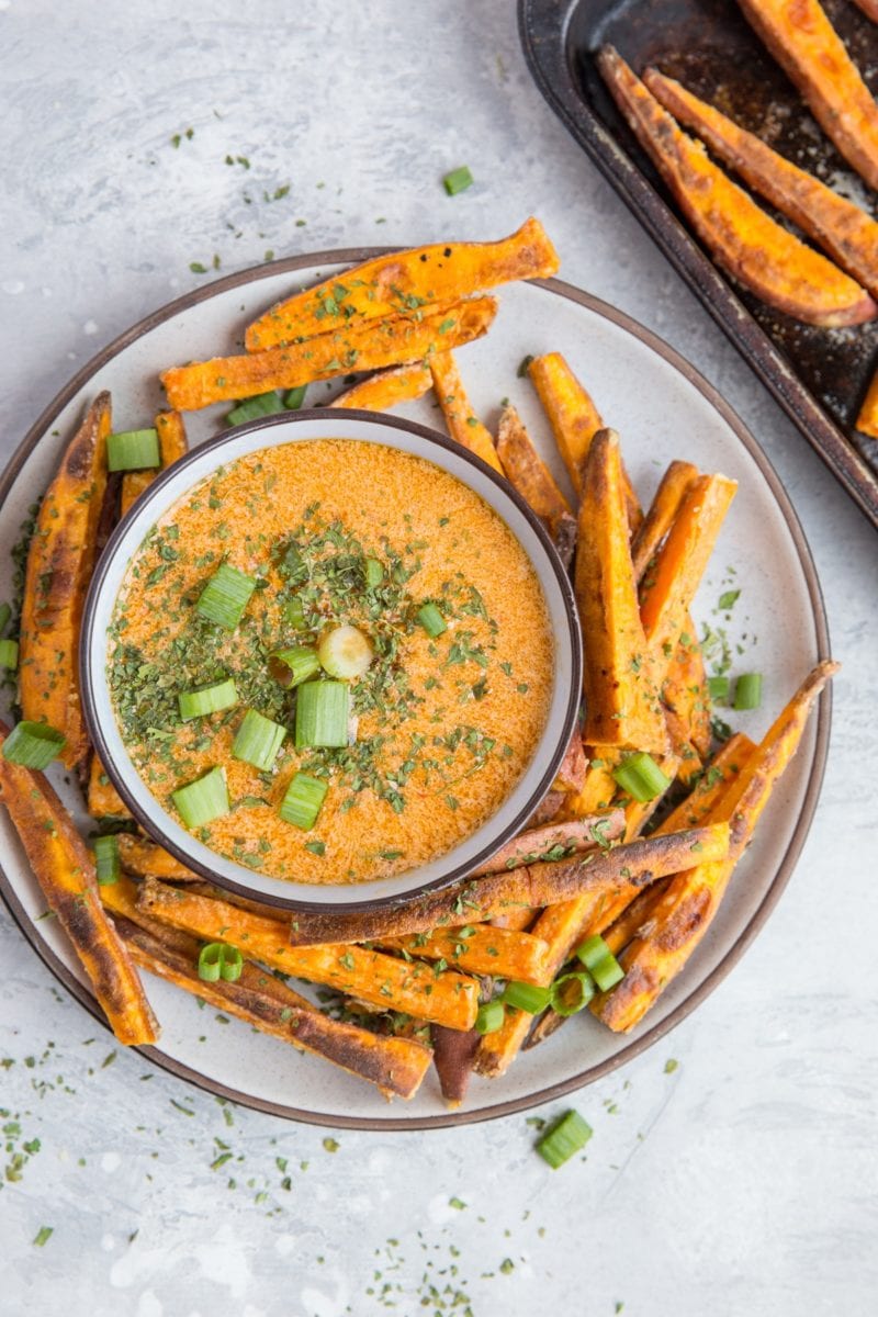 Sweet potato fries on a plate with a bowl of chipotle dipping sauce in the center, sprinkled with green onions. Ready to serve.