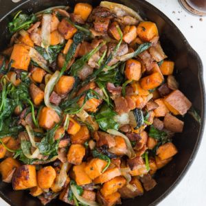 Sweet Potato Hash Recipe in a skillet with crispy bacon, onions, and spinach and a linen wrapped around the handle of the skillet. Pepper grinder to the side.