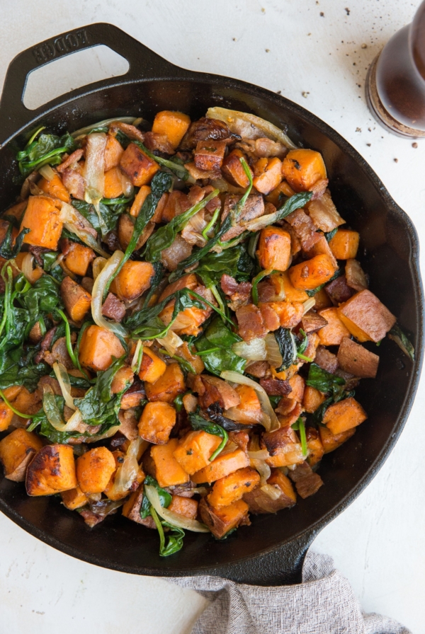 Sweet Potato Hash Recipe in a skillet with crispy bacon, onions, and spinach and a linen wrapped around the handle of the skillet. Pepper grinder to the side.