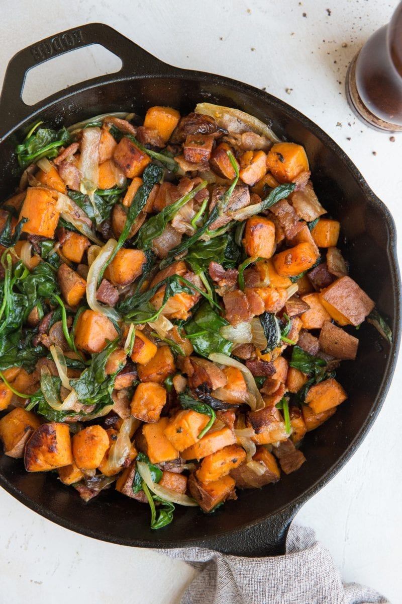 Sweet Potato Hash Recipe in a skillet with crispy bacon, onions, and spinach and a linen wrapped around the handle of the skillet. Pepper grinder to the side.