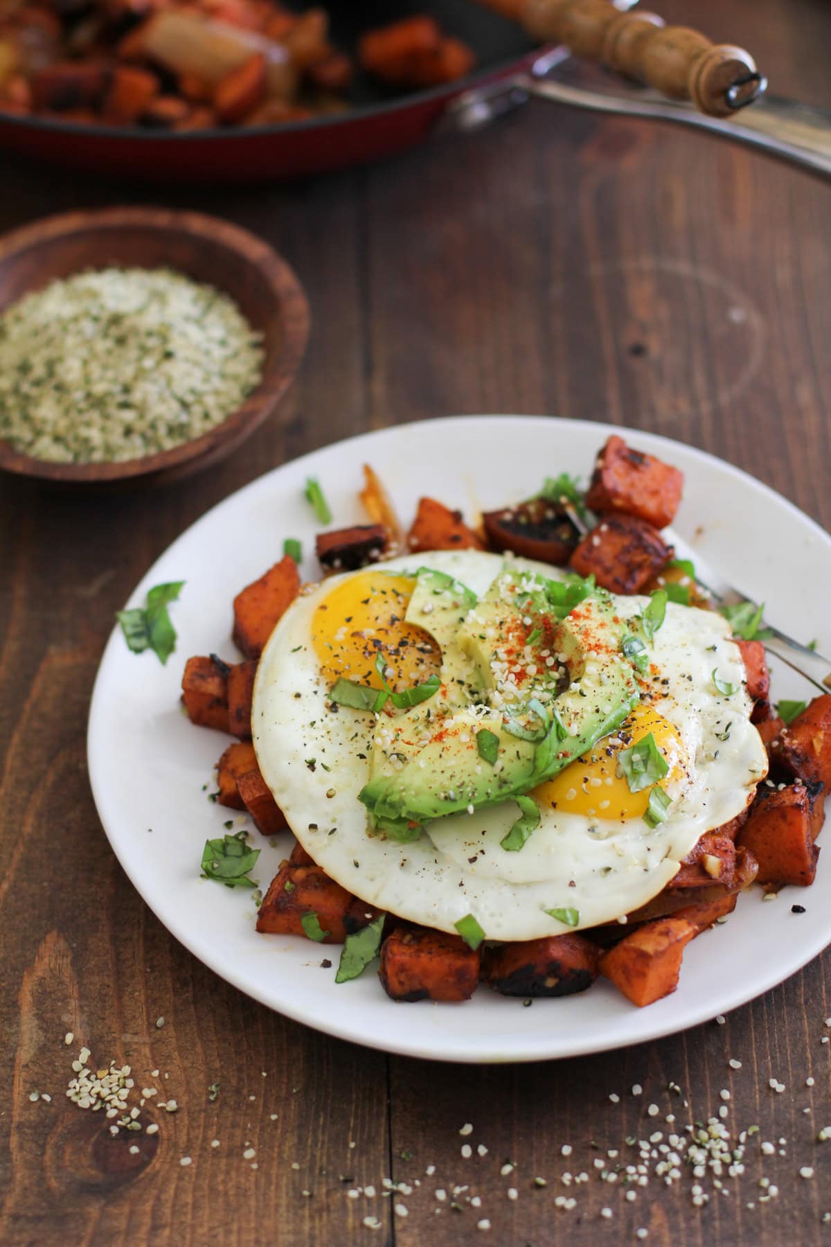 plate of sweet potato home fries with two fried eggs on top and sliced avocado, sprinkled with seasonings. The rest of the skillet in the background