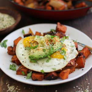 Small white plate with sweet potato home fries with fried eggs on top and sliced avocado, sprinkled with seasonings.