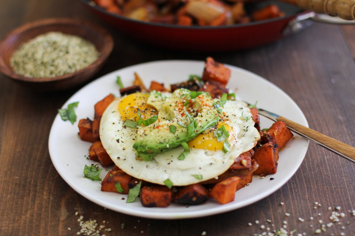 Small white plate with sweet potato home fries with fried eggs on top and sliced avocado, sprinkled with seasonings.