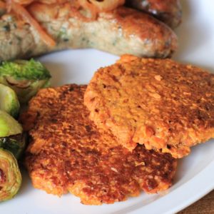 Rosti (hash browns) made with Sweet Potato on a white plate next to sauteed brussel sprouts and two sausages.