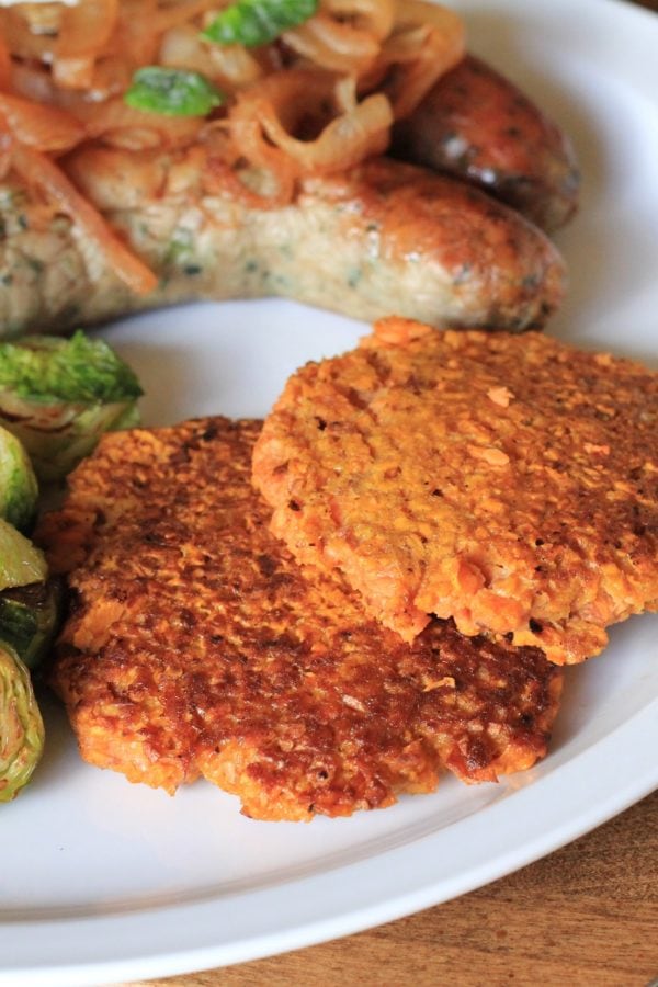 Rosti (hash browns) made with Sweet Potato on a white plate next to sauteed brussel sprouts and two sausages.