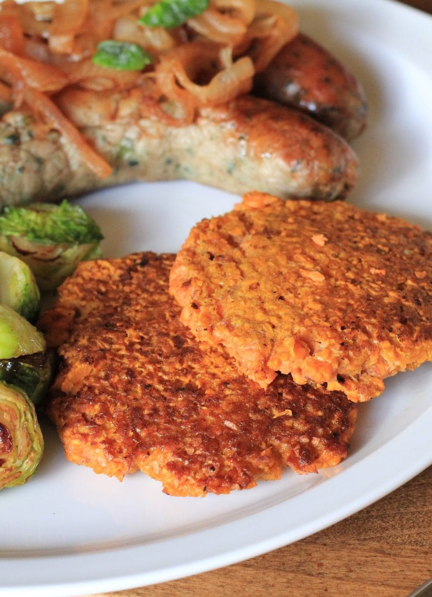 Rosti (hash browns) made with Sweet Potato on a white plate next to sauteed brussel sprouts and two sausages.
