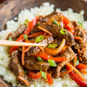 Szechuan beef in a large wooden bowl on top of cauliflower rice with a pair of chopsticks taking a bite of beef.