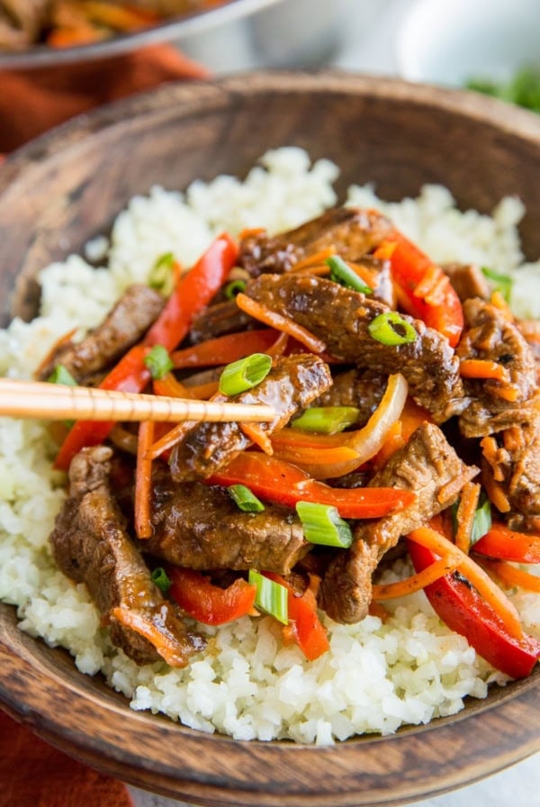 Szechuan beef in a large wooden bowl on top of cauliflower rice with a pair of chopsticks taking a bite of beef.