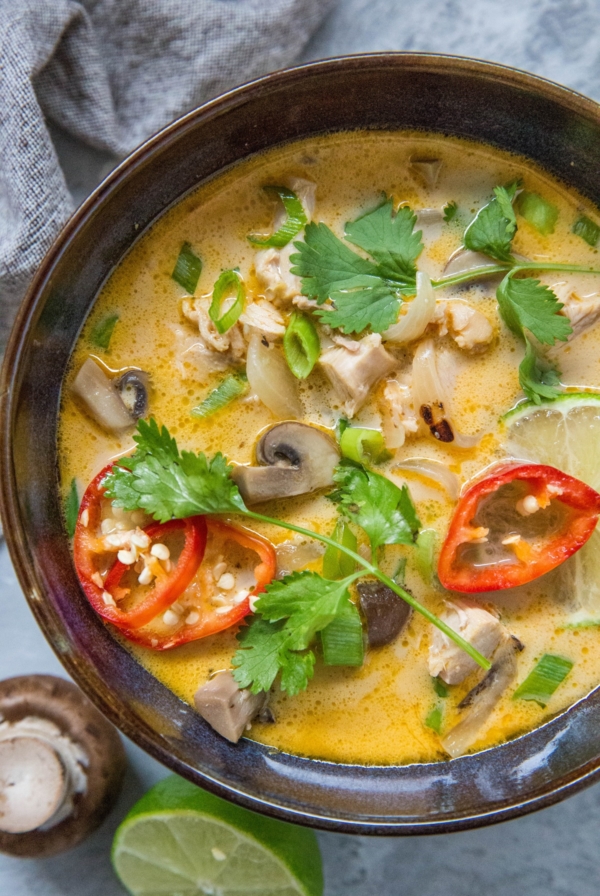Tom kha gai soup in a big bowl, ready to serve. A fresh mushroom and a half of a lime next to the bowl of soup.