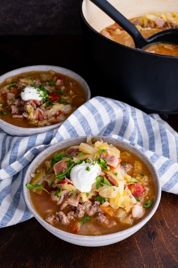 Sausage and cabbage stew in two bowls with a pot of soup in the background