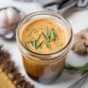 Tri Tip marinade in a jar with fresh rosemary on top of the marinade, a bulb of fresh garlic to the side and a whisk.