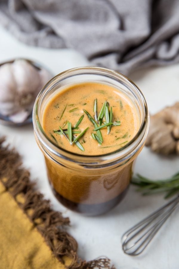Tri Tip marinade in a jar with fresh rosemary on top of the marinade, a bulb of fresh garlic to the side and a whisk.