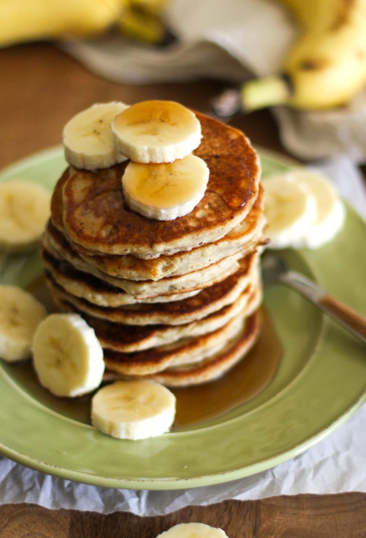 Silver Dollar Vegan Banana Pancakes on a green plate drizzled with pure maple syrup and fresh sliced bananas on top.