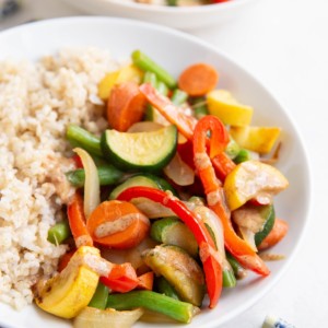 Big white bowl of vegetable stir fry with peanut sauce and brown rice with an orange napkin and blue chopsticks to the side.