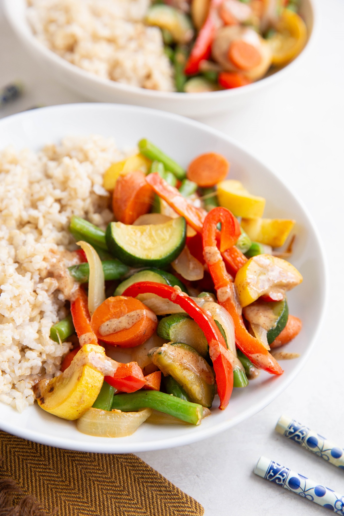 Big white bowl of vegetable stir fry with peanut sauce and brown rice with an orange napkin and blue chopsticks to the side.