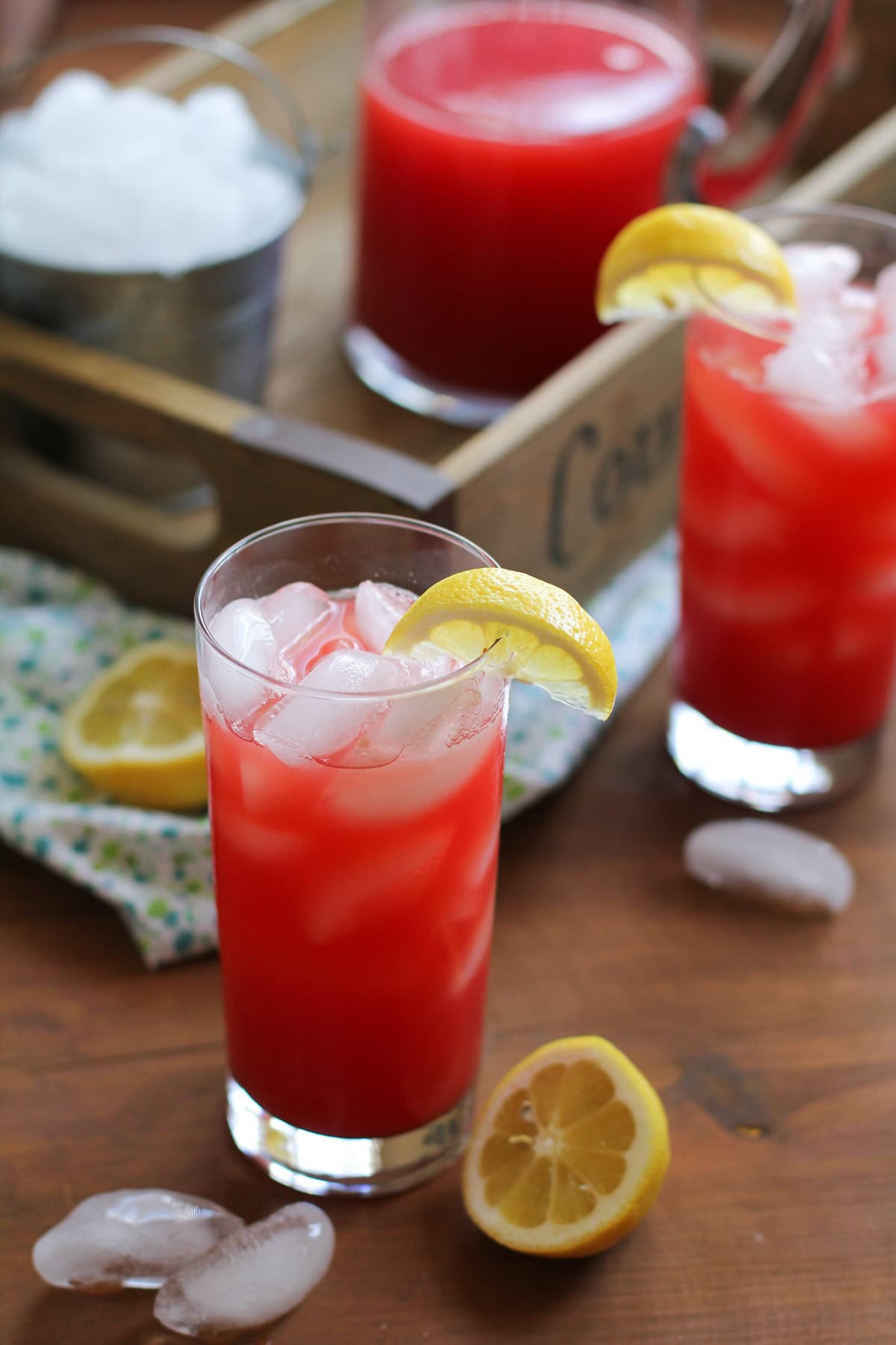 A glass of Agua Fresca sitting on a wooden table with a slice of lemon around the rim, a half a lemon to the side, and another glass and pitcher of agua fresca in the background. Ready to serve.