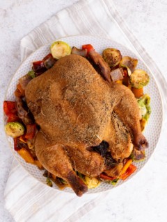 Whole smoked chicken on a plate on top of roasted vegetables, fresh off the smoker grill and ready to serve.