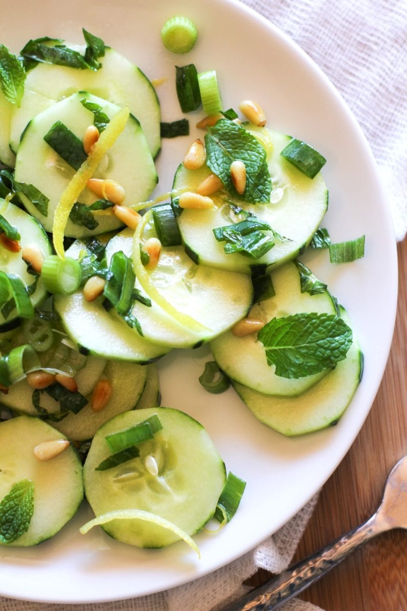 Zesty Cucumber Salad with Pine Nuts on a white plate with green onions and mint leaves and a fork to the side.