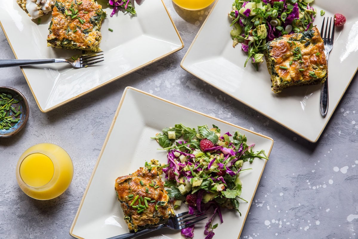 Three plates with slices of breakfast casserole with salad and mimosas to the side for a delicious brunch.