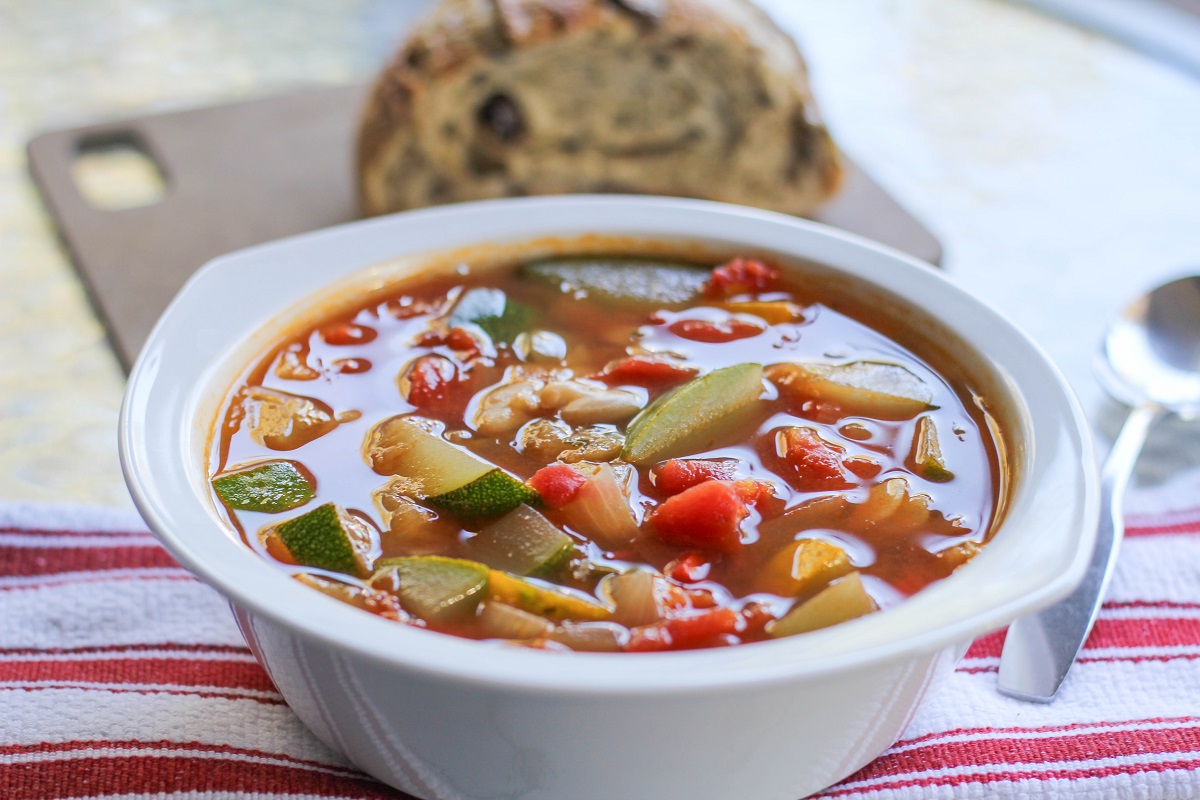 White bowl of zucchini and yellow squash soup with beans and a napkin and spoon to the side.