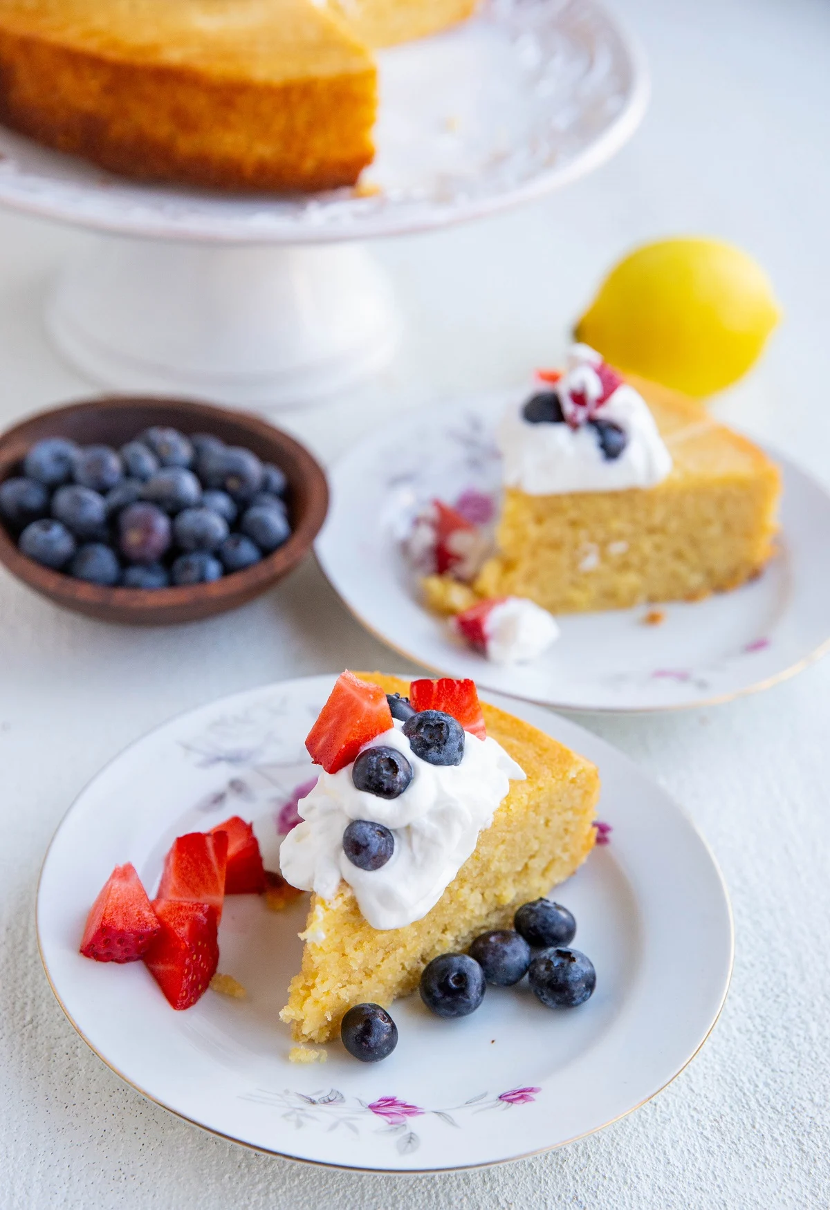 Two slices of lemon cake on plates with whipped cream on top and berries.