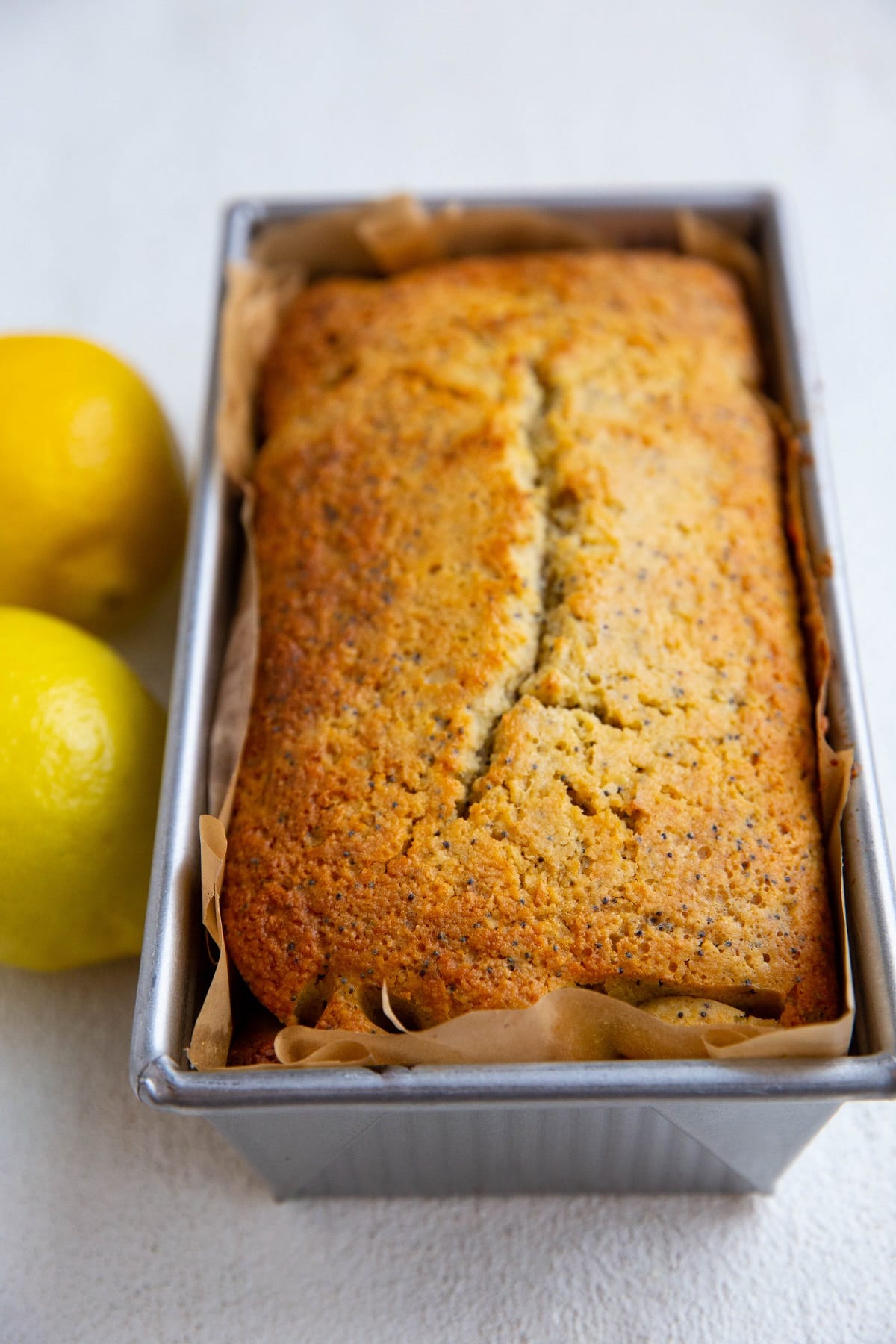 Loaf of lemon poppy seed bread in a bread pan fresh out of the oven with fresh lemons to the side.