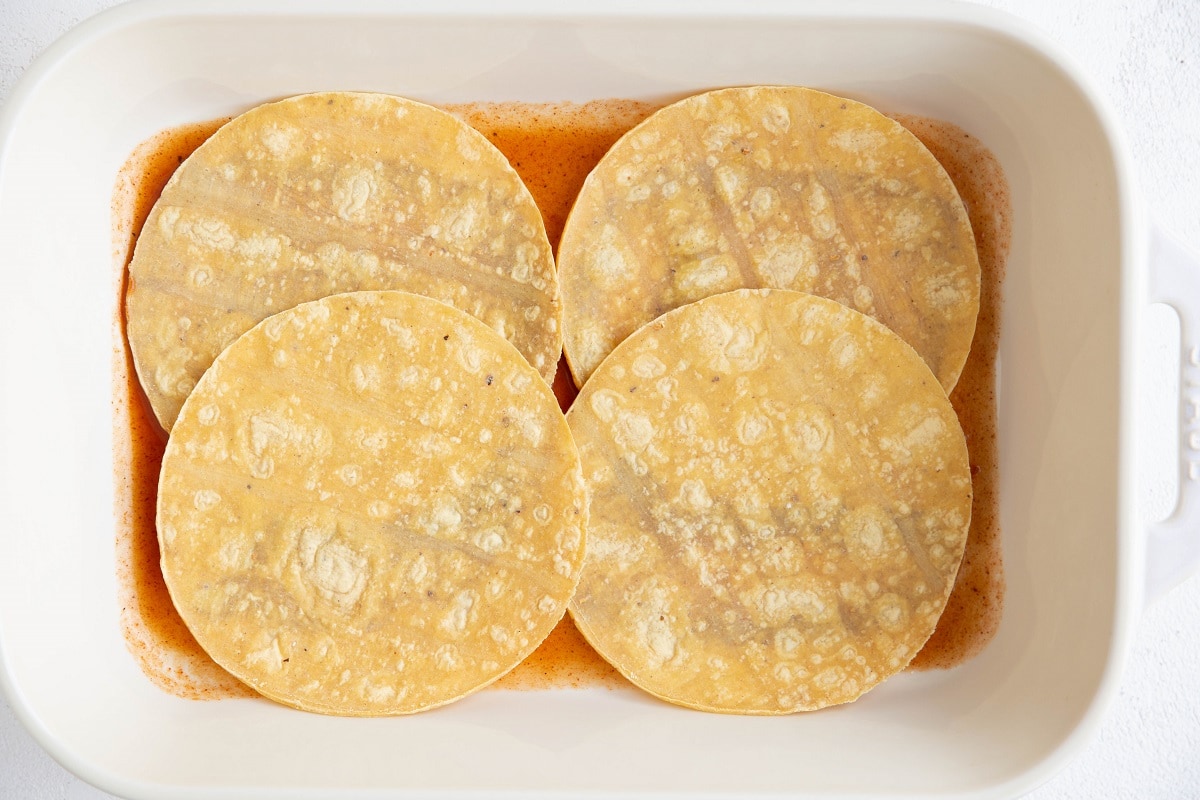 Corn tortillas on top of enchilada sauce in a baking dish.