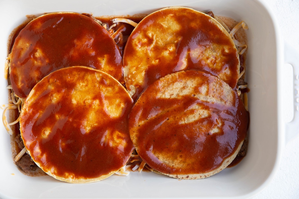 Corn tortillas creating a second layer for enchilada lasagna with enchilada sauce spread on top.