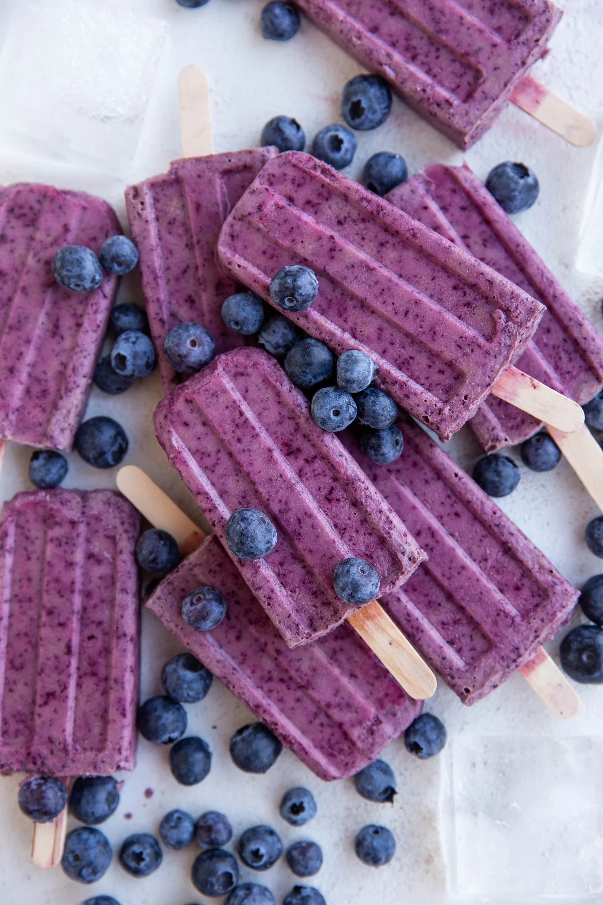 Pile of blueberry coconut milk popsicles on a white background with ice and fresh blueberries all around.