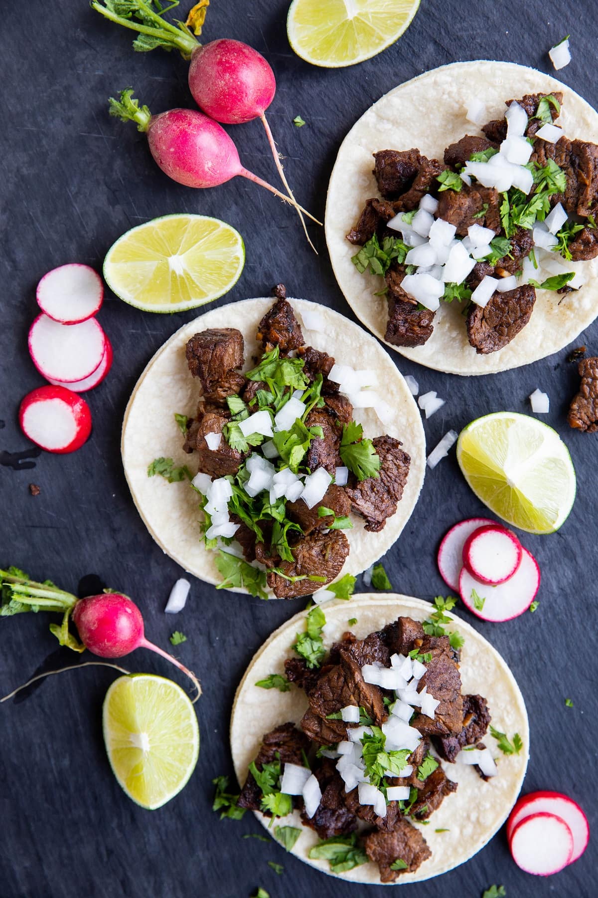 Large black serving platter with carne asada tacos on top. Sliced radishes and lemon wedges to the side.