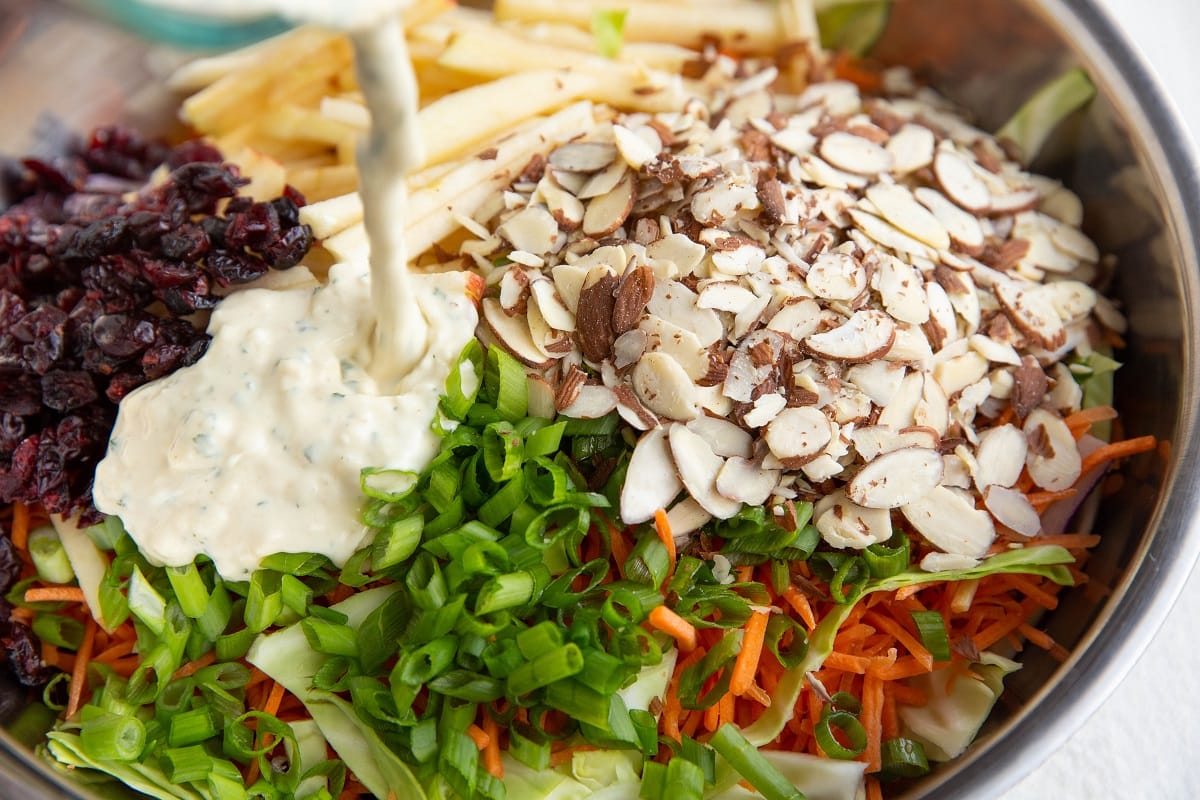 Mixing bowl with veggies and ingredients for coleslaw with dressing being poured in.