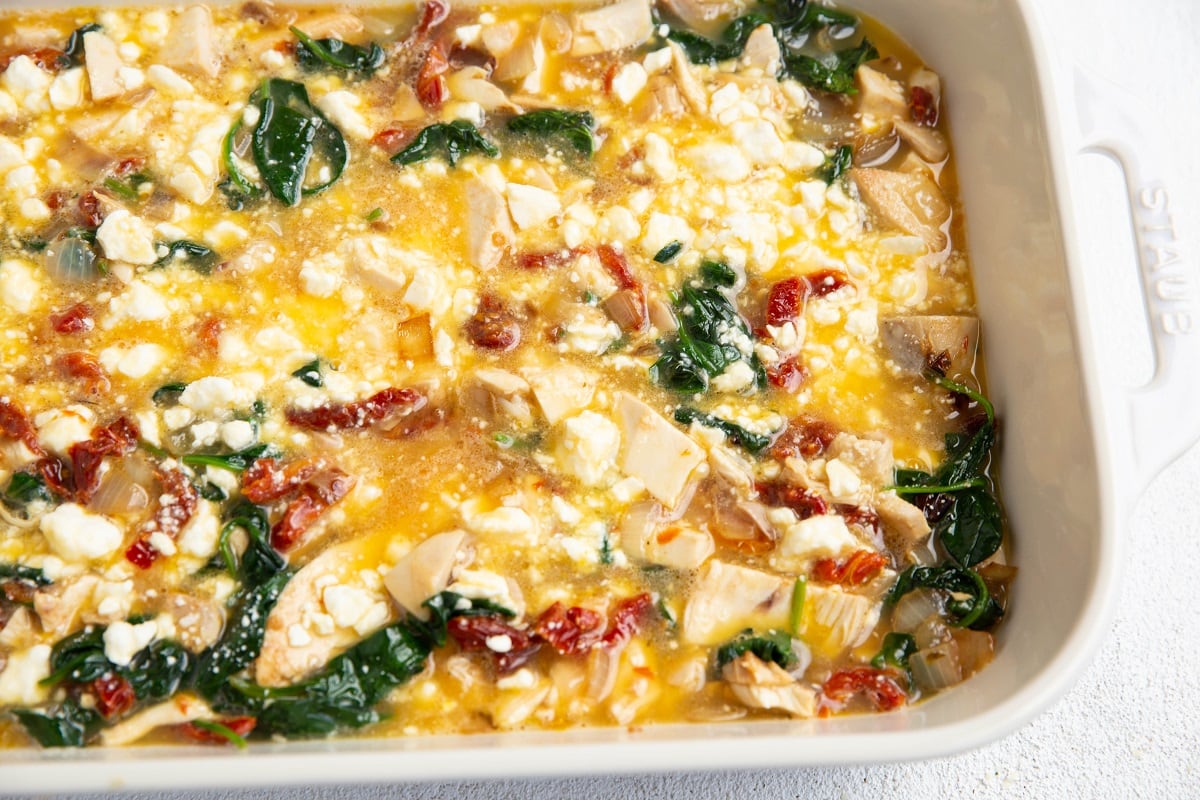 Feta cheese stirred into raw eggs, vegetables and chicken in a casserole dish, ready to bake.