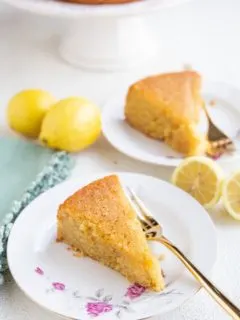 Two slices of lemon cake on plates with the rest of the cake in the background and fresh lemons all around.
