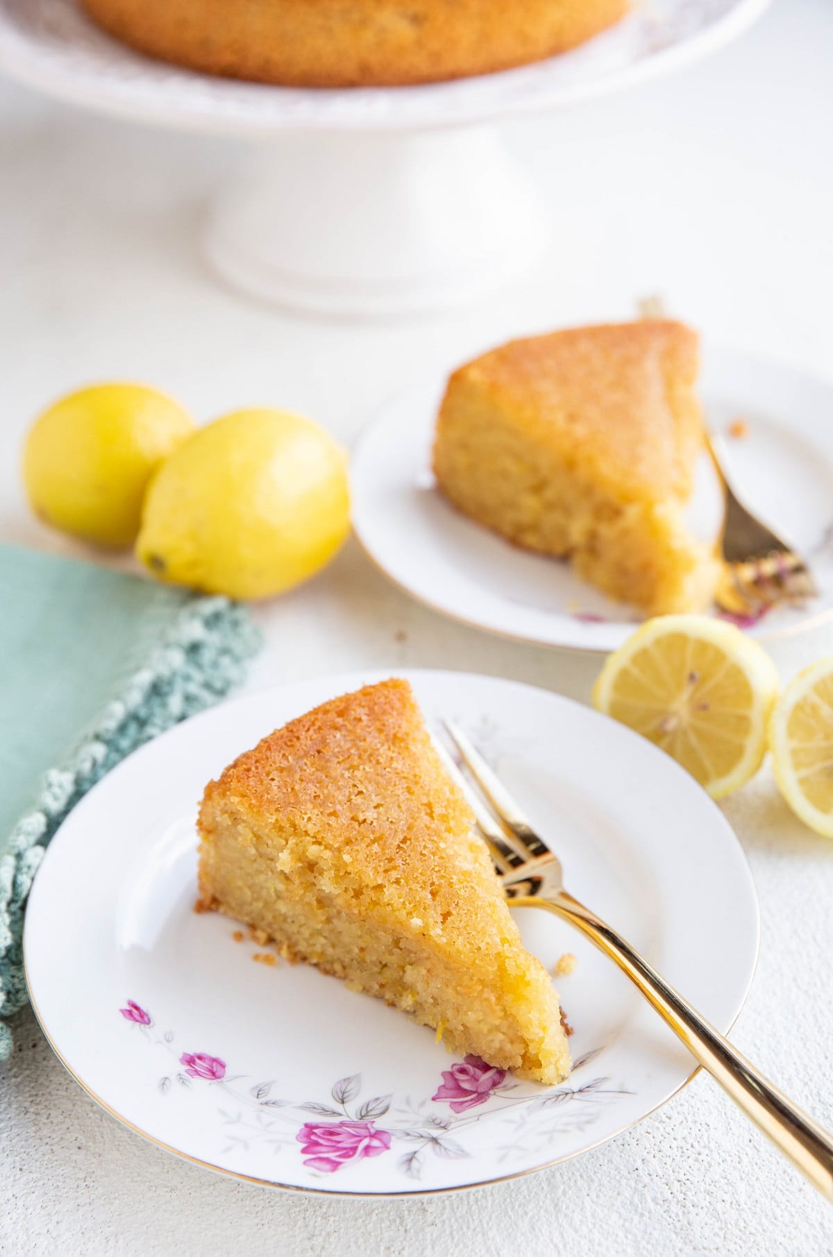 Two slices of lemon cake on plates with the rest of the cake in the background and fresh lemons all around.
