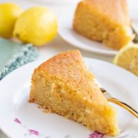 Two slices of lemon cake on two plates with fresh lemons all around.