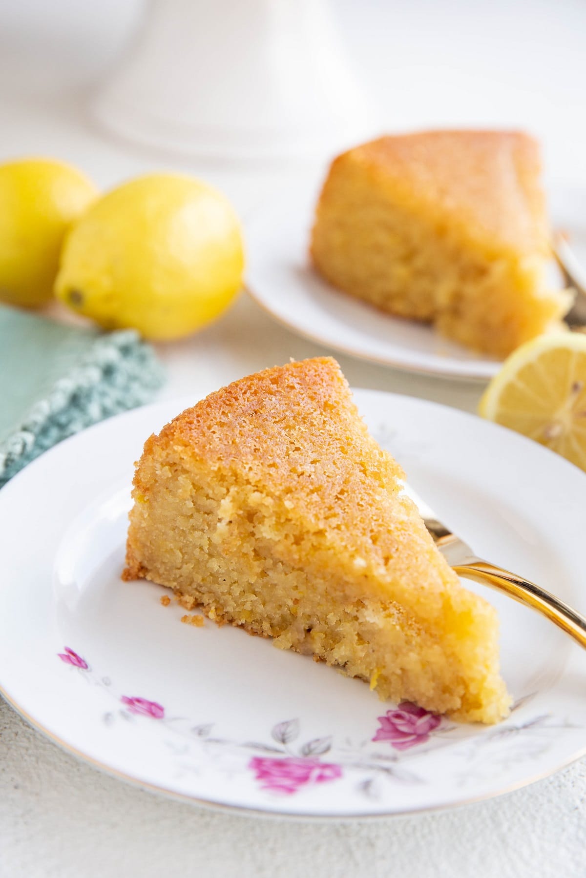 Two slices of lemon cake on two plates with fresh lemons all around.