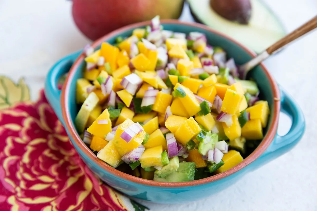 Bowl of mango salsa with a napkin to the side and juicy mango and ripe avocado in the background.