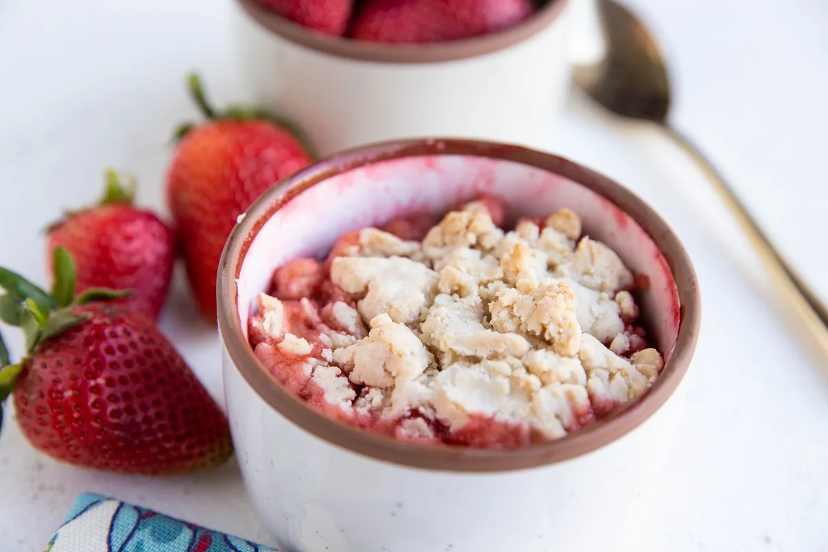 White ramekin of strawberry crumble with fresh strawberries to the side, ready to serve.