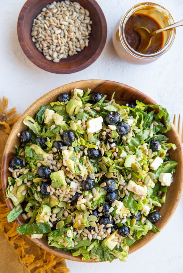 Bowl of green salad with blueberries, avocado, sunflower seeds, feta cheese, pumpkin seeds and balsamic vinaigrette mixed in.