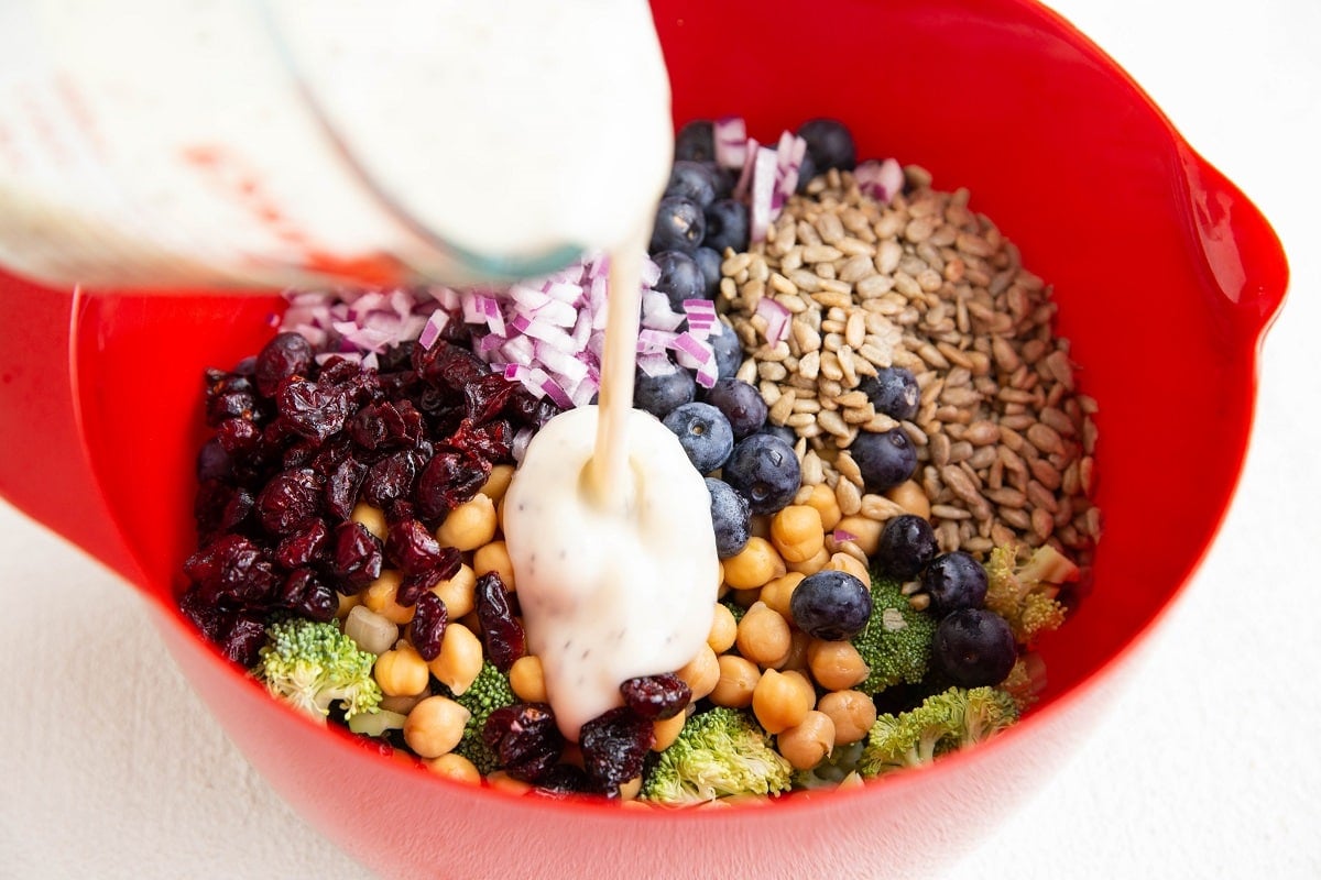 Pouring salad dressing over the broccoli salad ingredients in a mixing bowl.