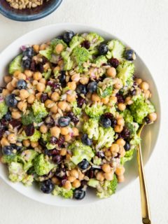 Big white bowl of broccoli chickpea salad with a gold spoon and a bowl of sunflower seeds.