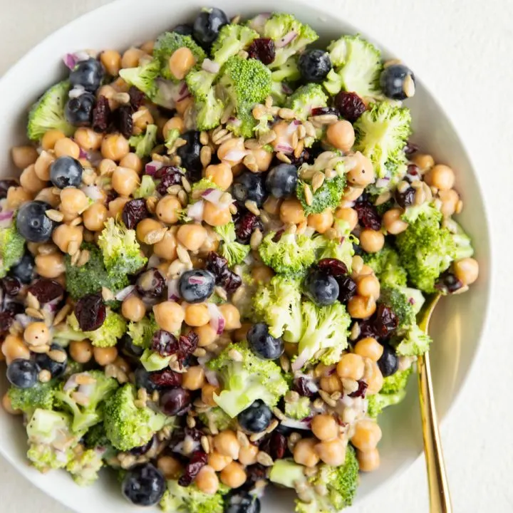 Big white bowl of broccoli chickpea salad with a gold spoon and a bowl of sunflower seeds.